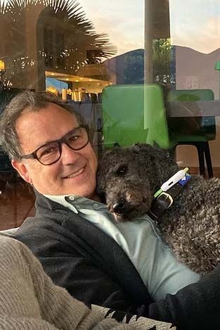 Ein Mann mit Brille lächelt und kuschelt mit einem schwarzen Hund; er sitzt drinnen und hat einen Fensterblick.