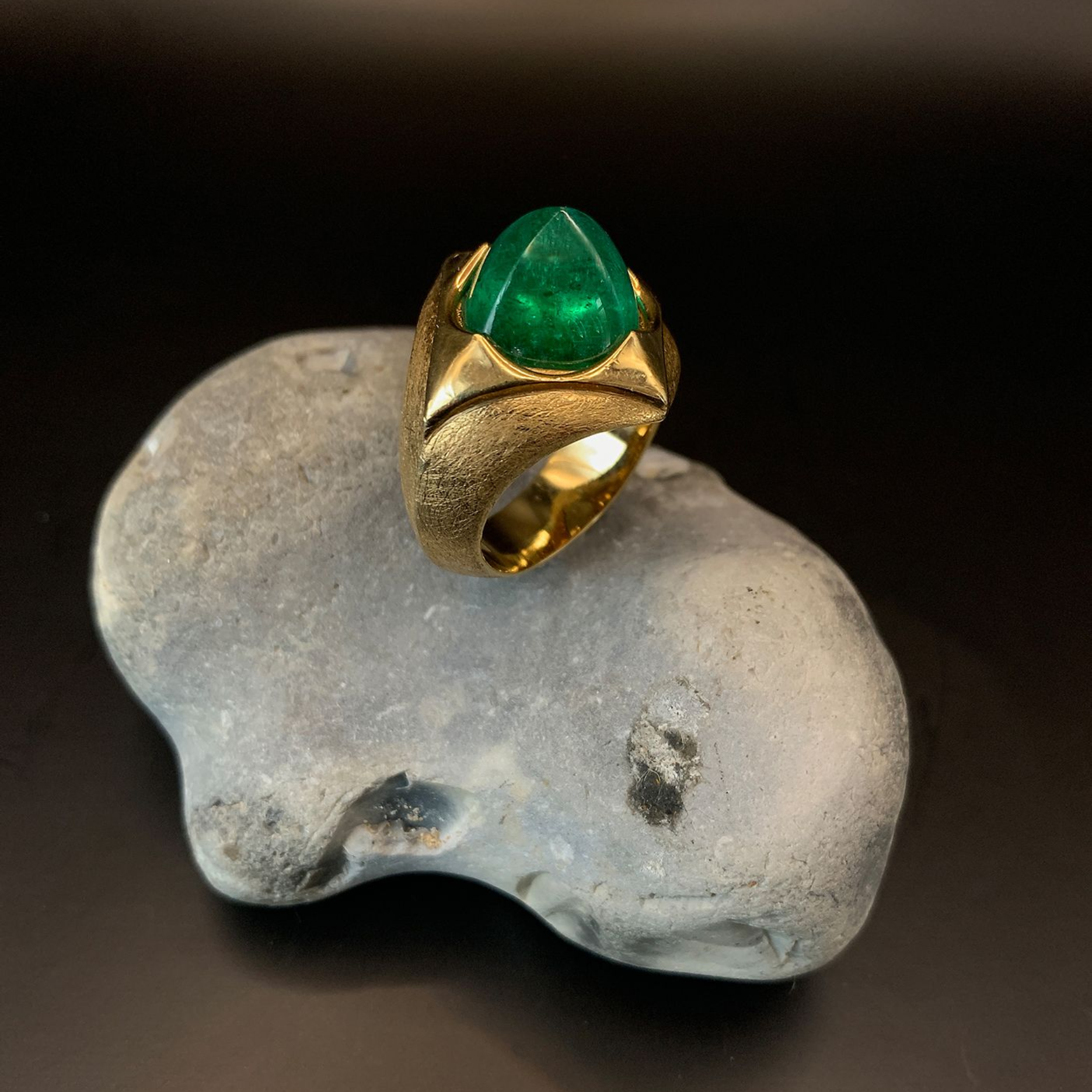 Goldring mit einem grünen Edelstein auf grauem Grund vor dunklem Hintergrund