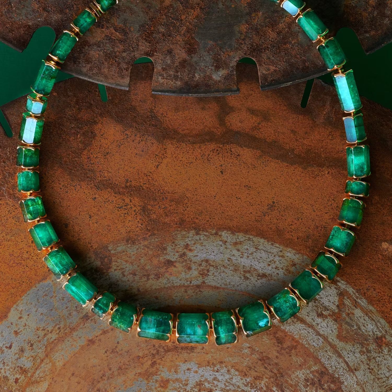 Eine Smaragdkette mit Goldfassung hebt sich von einer rostbraunen Metalloberfläche ab.