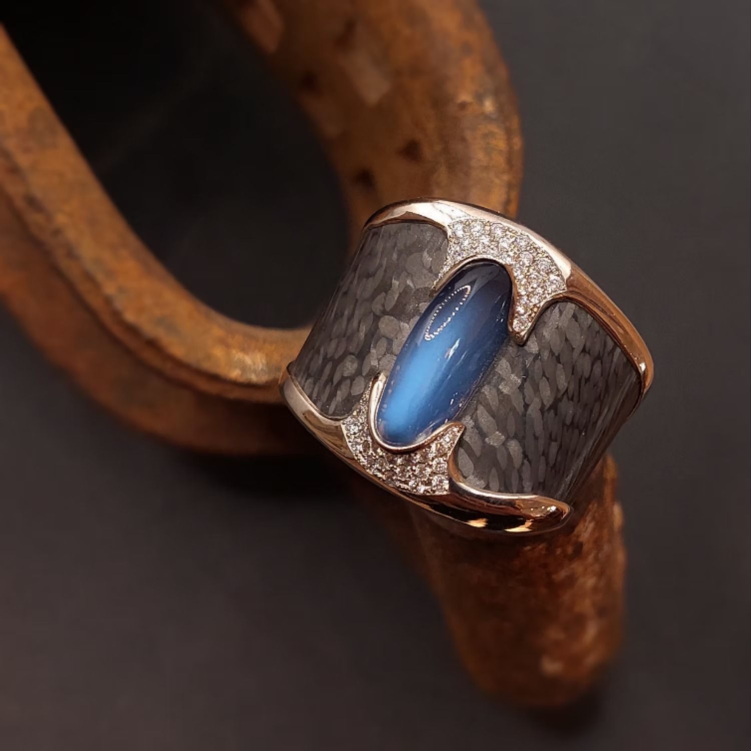 Ein Ring aus Roségold mit einem blauen Edelstein, Kohlefaser-Einlage und Diamantakzenten, ruhend auf einem rostigen Hufeisen.