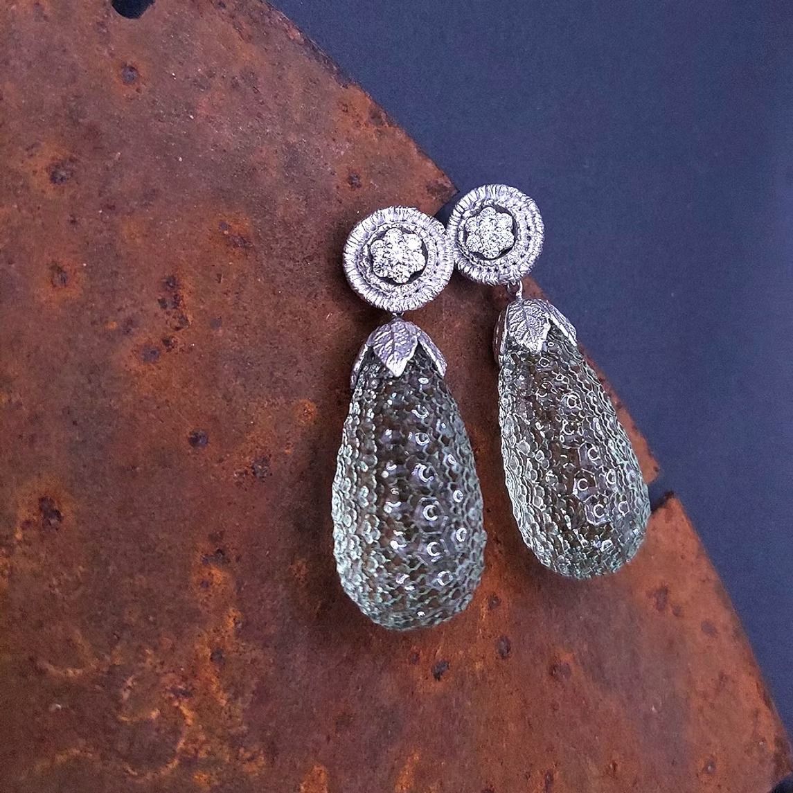 Ohrhänger mit Diamanten und grünen Edelsteinen auf einer strukturierten, rostbraunen Oberfläche.