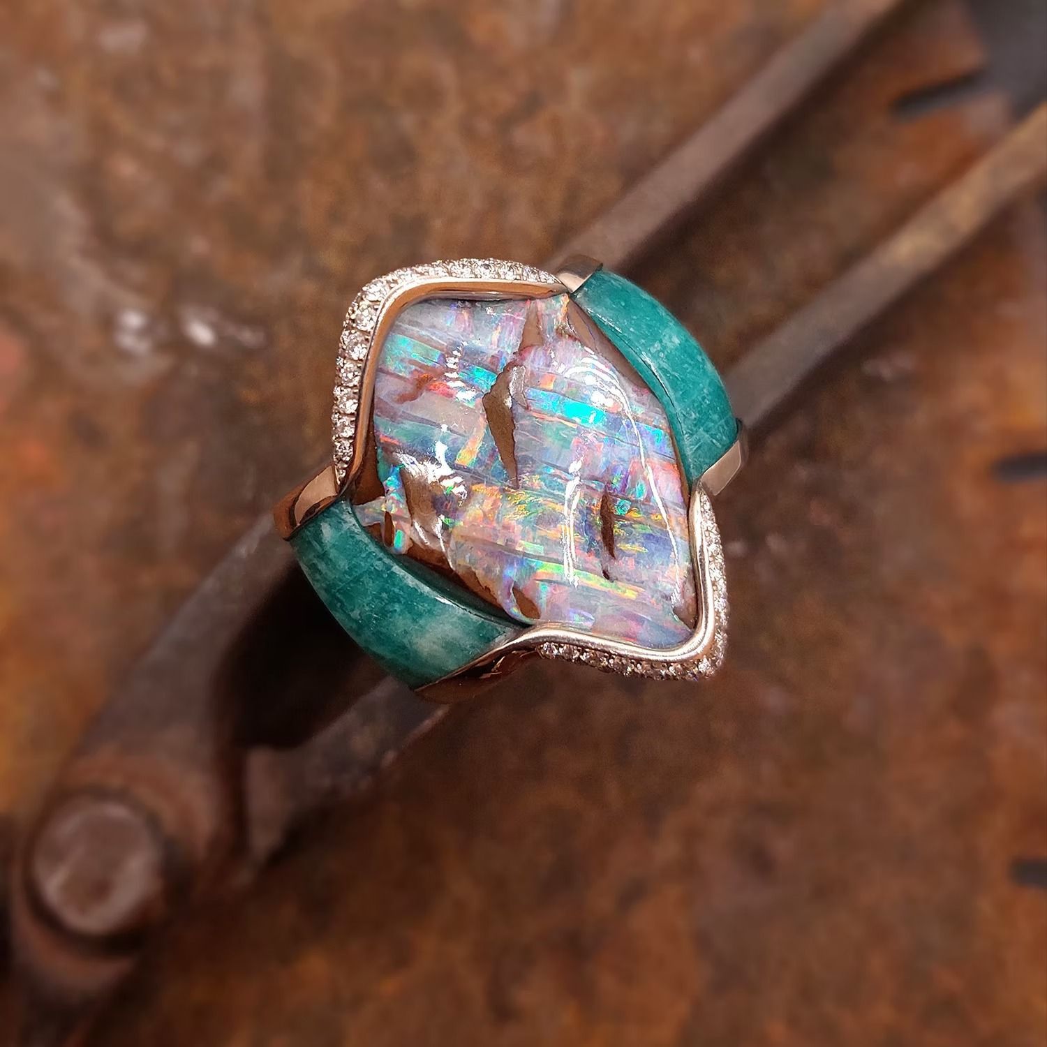 Ein Ring aus Roségold mit einem großen Opal, türkisfarbenen Akzenten und kleinen Diamanten, der auf Metallwerkzeugen ruht.