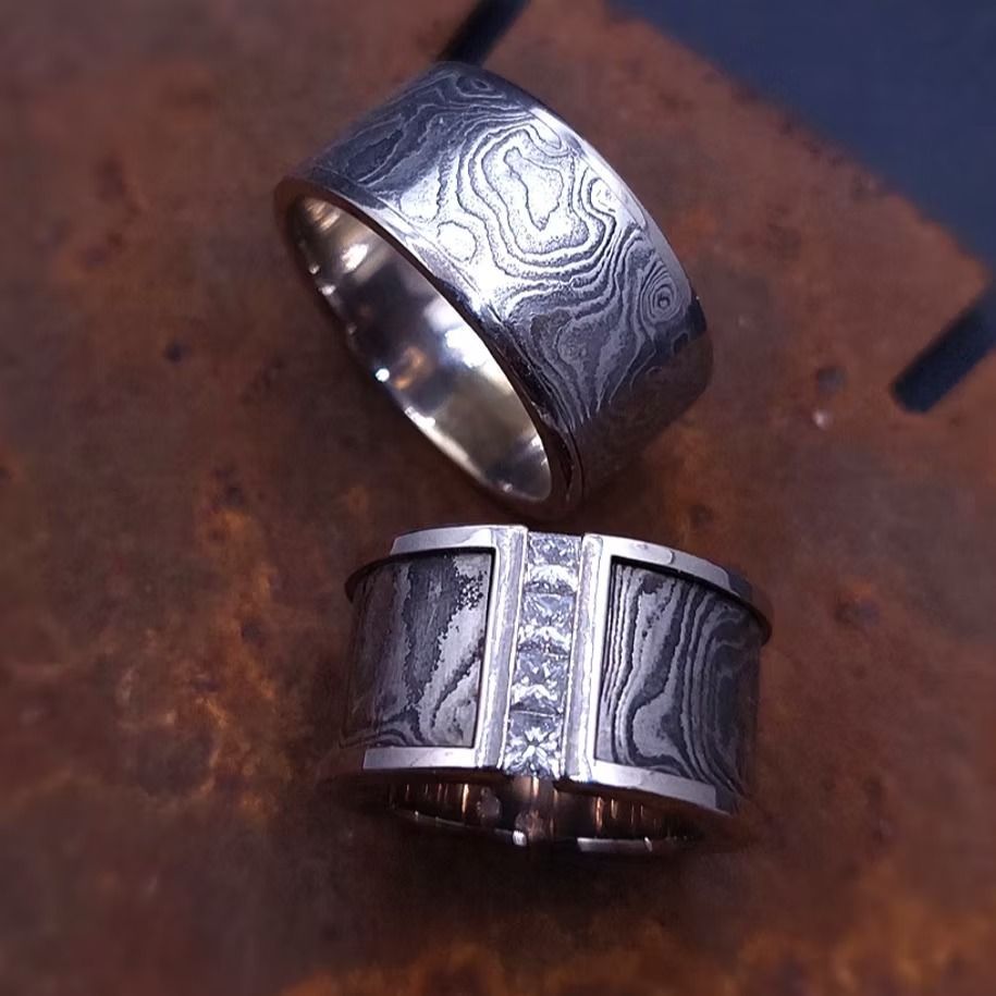 Zwei Eheringe mit strukturierten, blaugrauen Einlagen, wobei ein Ring auf dem anderen ruht, auf einer dunklen Oberfläche.