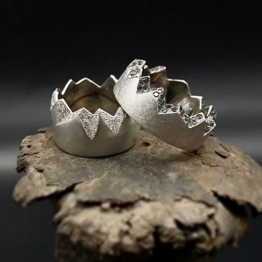 Zwei silberne Ringe mit gezackten Kanten, besetzt mit kleinen Steinen, ruhen auf einer strukturierten, dunklen Oberfläche.