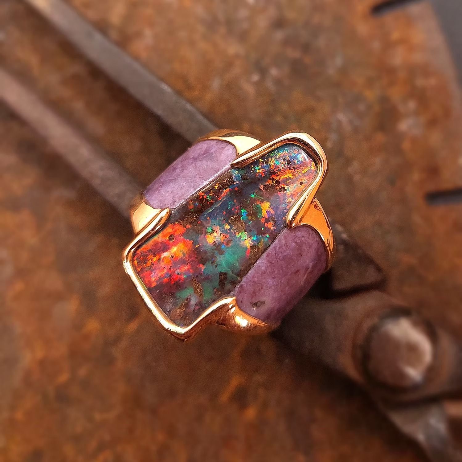 Goldring mit schillerndem Opal und violetten Steinen auf rostiger Metalloberfläche