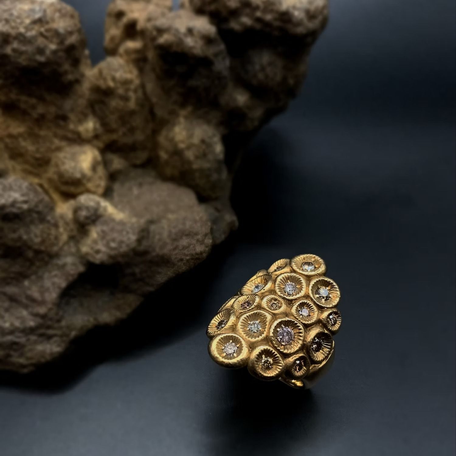 Ein goldener Ring mit Diamantakzenten, der neben einem strukturierten braunen Stein auf dunklem Hintergrund präsentiert wird.