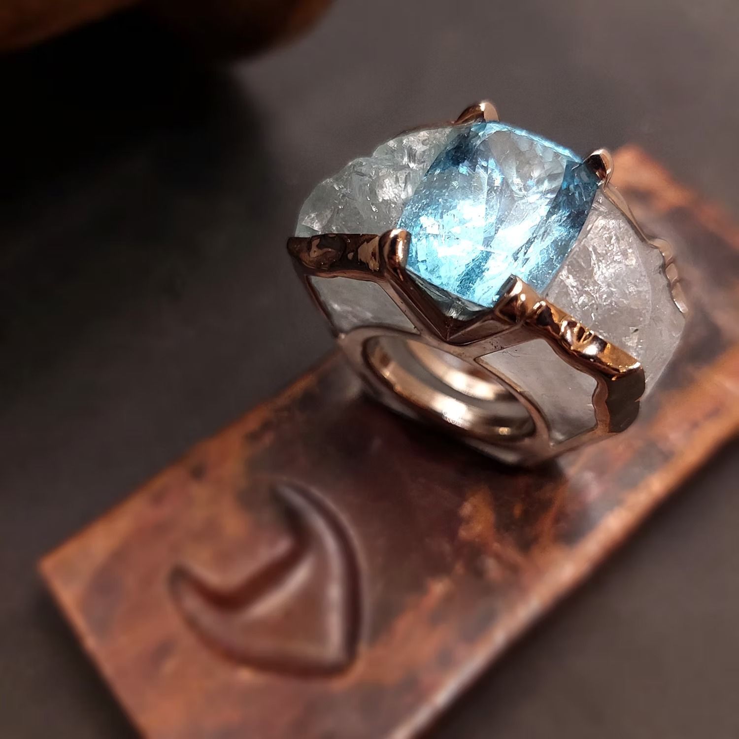 Ring mit einem großen, grob geschliffenen blauen Edelstein und weißen Seitensteinen, gefasst in Roségold, auf einer strukturierten Bronzeoberfläche.