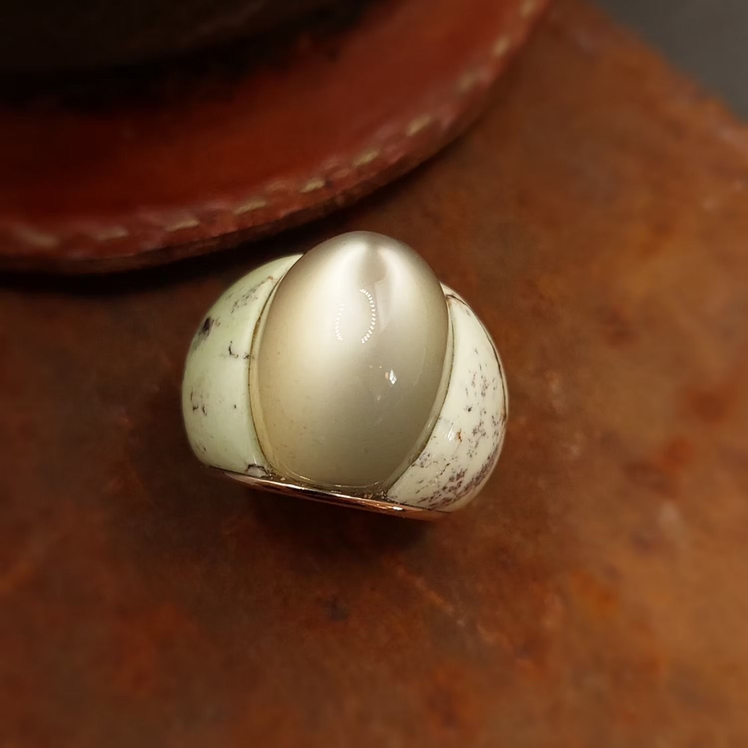 Ovaler Ring mit einem großen perlmuttartigen Stein, der zwischen hellgrünen und gesprenkelten weißen Seiten auf braunem Grund eingefasst ist.