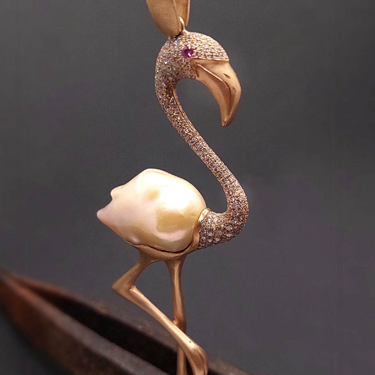 Goldener Flamingo-Anhänger mit Perlenkörper, diamantbesetztem Hals und Rubinauge, gehalten von einer Pinzette.
