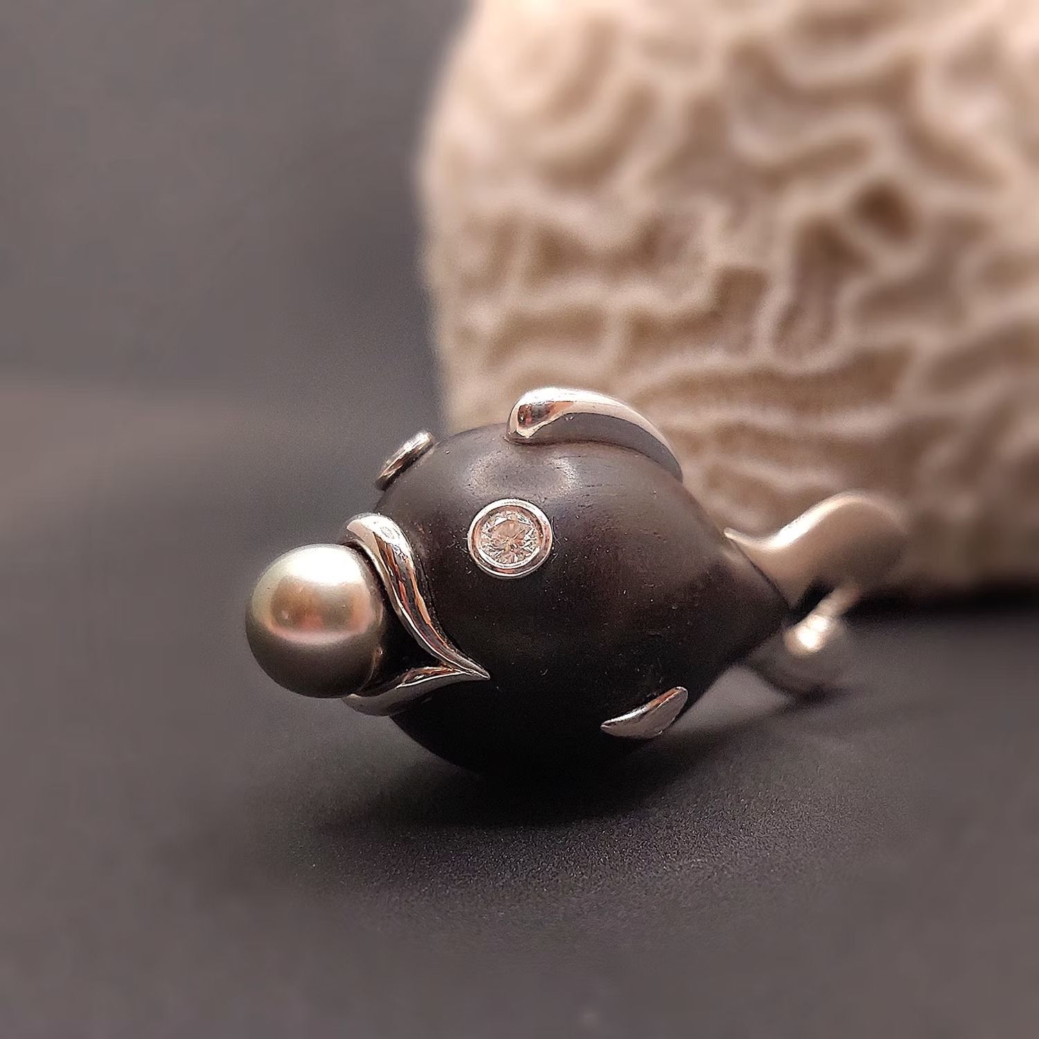 Dunkle Fischbrosche mit Perlen- und Diamantverzierungen vor strukturiertem Korallenhintergrund.
