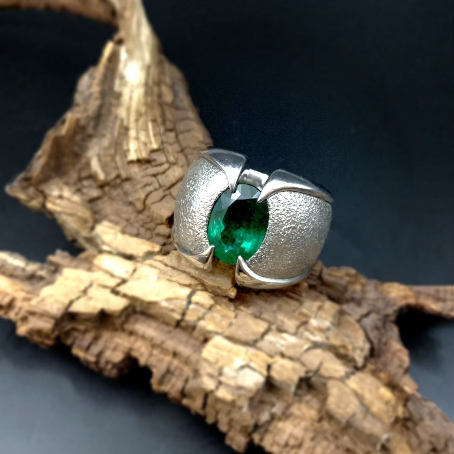 Silberner Ring mit einem grünen Edelstein auf einem Stück Rinde vor dunklem Hintergrund