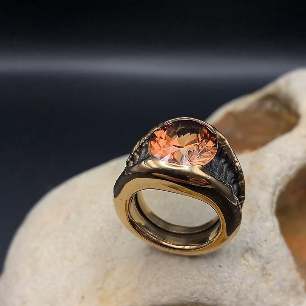 Nahaufnahme eines Goldrings mit einem dunklen, orange gesprenkelten Stein auf hellem Untergrund