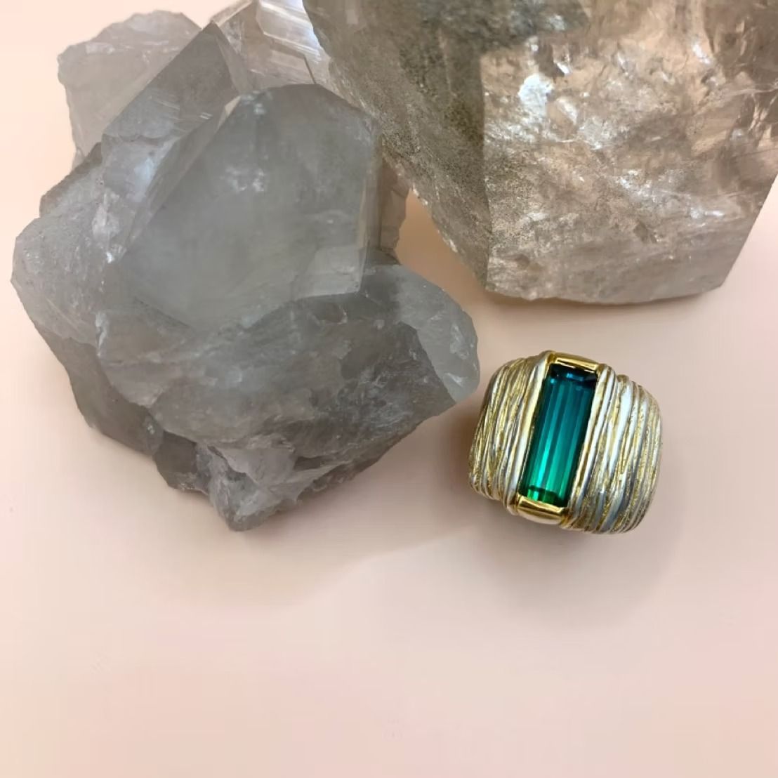 Ring mit rechteckigem grünem Edelstein, goldenen Akzenten, neben Quarzkristallen auf rosafarbenem Grund.