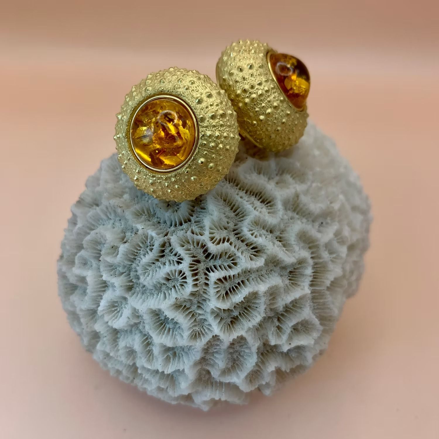 Goldfarbene Ohrringe mit bernsteinfarbenen Mittelsteinen ruhen auf einer porösen, weißen Korallenformation vor einem rosa Hintergrund.