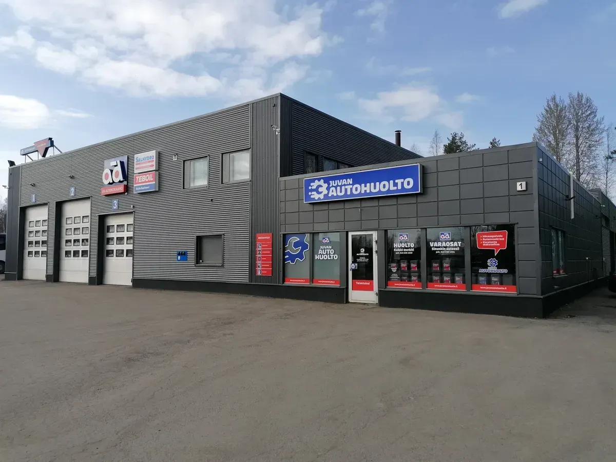 Moderni tummanharmaa autokorjaamo, jossa on autotallipaikkoja, punaisia yksityiskohtia ja sininen kyltti pilvisen taivaan alla