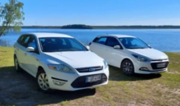 Kaksi valkoista autoa pysäköitynä nurmikolle järven rannalle kirkkaan taivaan alle