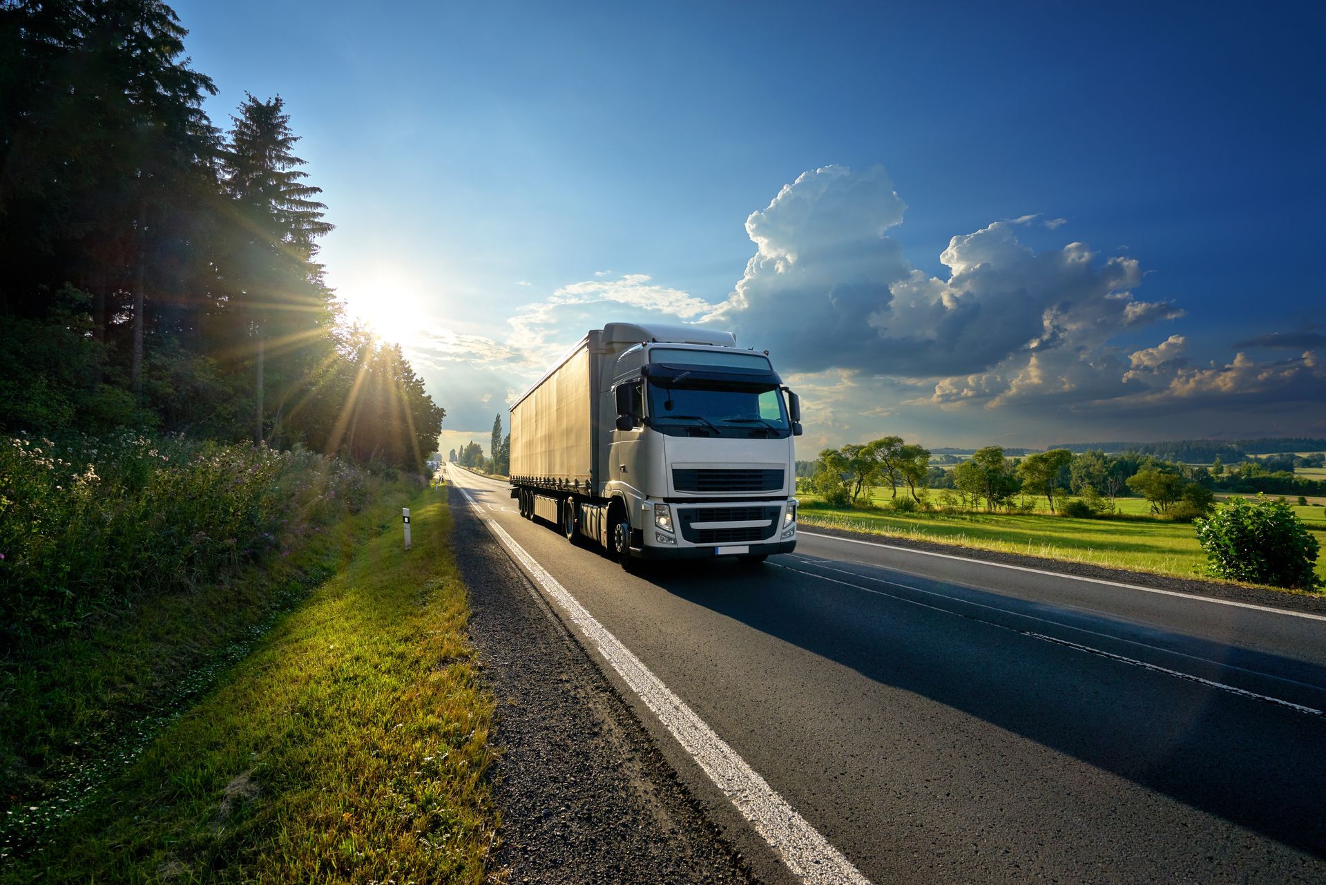 Un camion roule sur une route ensoleillée, bordée de champs verdoyants et d'arbres au coucher du soleil.