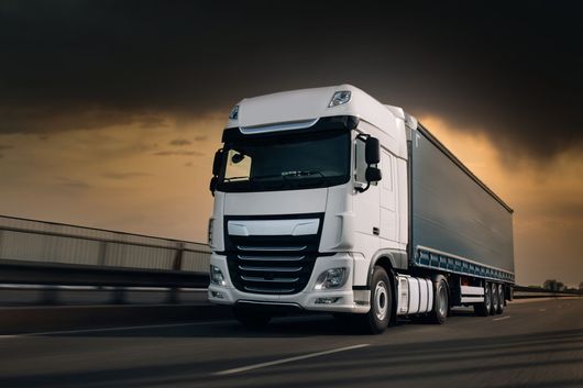 Un semi-remorque blanc circule sur une autoroute au crépuscule, sous un ciel nuageux.