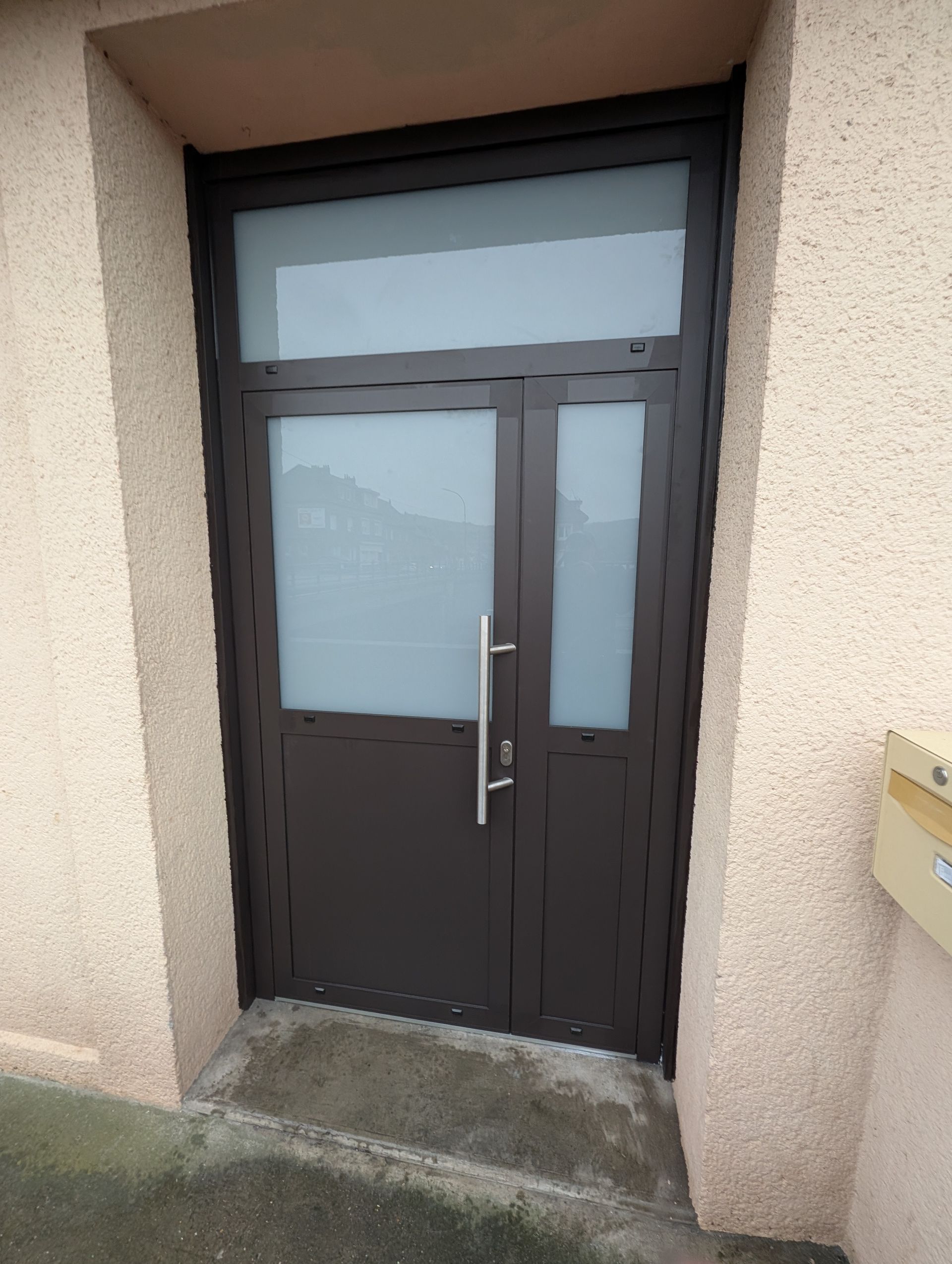 Porte marron avec panneaux de verre dépoli, encastrée dans un mur extérieur beige, poignée en métal.