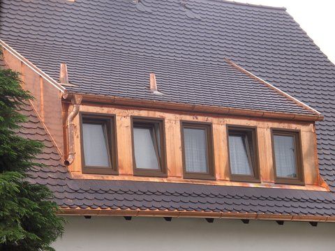 Ein mit grauen Schindeln gedecktes Dach mit Giebelgaube, blauer und hellgrauer Verkleidung und einem zentralen Fenster vor blauem Himmel.