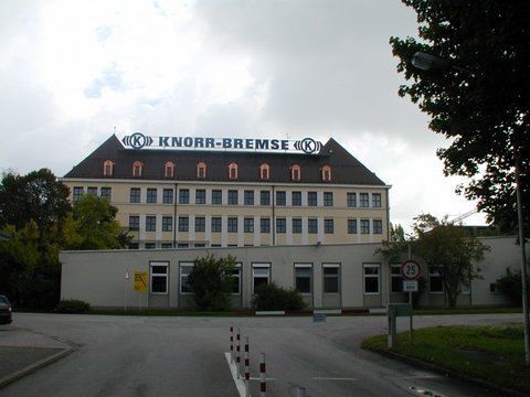 Ein großes Fabrikgebäude von Knorr-Bremse mit einem Schild auf dem Dach, gesehen von einer Asphaltstraße unter bewölktem Himmel.