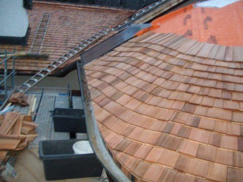 Eine Draufsicht auf ein im Bau befindliches Holzschindeldach mit orangefarbener Unterspannbahn und einem nahegelegenen Gerüst.