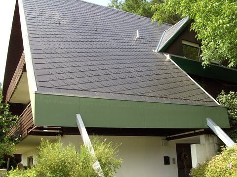 Ein Haus mit steilem, dunkel geschindeltem Dach, grüner Fassade, weißen Wänden und schrägen Stützpfeilern, umgeben von Bäumen.