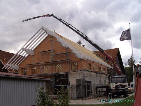 Eine Baustelle, auf der ein Kran unter bewölktem Himmel ein Dachsegment auf ein Backsteingebäude hebt.