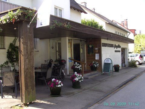 Ein sonniger Außenbereich mit Sitzgelegenheiten an einem Restaurantgebäude, mit blumengefüllten Töpfen und einer überdachten Terrasse.