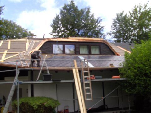 Ein Arbeiter repariert auf einem Gerüst das Dach eines Hauses mit Dachgaube, umgeben von Bäumen unter einem teilweise bewölkten Himmel.