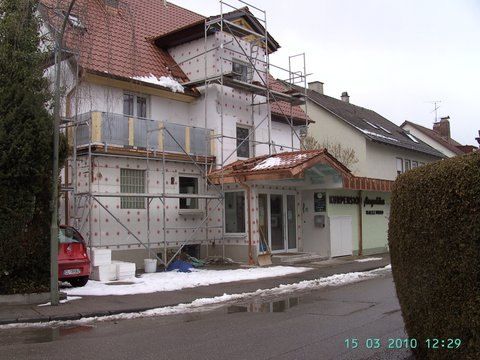 Ein Wohngebäude, dessen Fassade renoviert wird, ist auf einer verschneiten Straße mit Gerüsten und weißen Dämmplatten versehen.