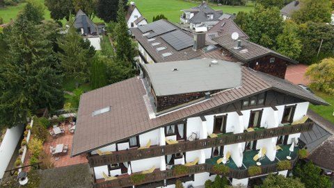 Luftaufnahme eines mehrstöckigen Hotels im alpinen Stil mit braunen Dächern, weißen Wänden und terrassenförmigen Balkonen inmitten grüner Bäume.