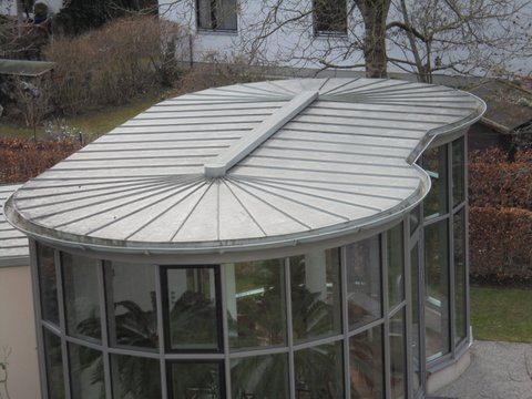 Ein nierenförmiges Metalldach mit strahlenförmigen Paneelen überspannt einen modernen, verglasten Wintergarten in einer Gartenanlage.