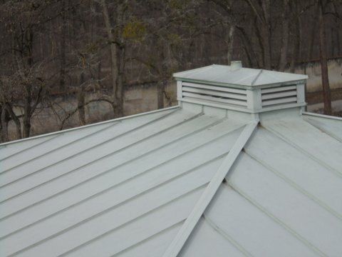 Ein helles Metalldach mit Stehfalzen und einer quadratischen, mit Lüftungsschlitzen versehenen Kuppel, die mittig auf dem Dachfirst angeordnet ist.