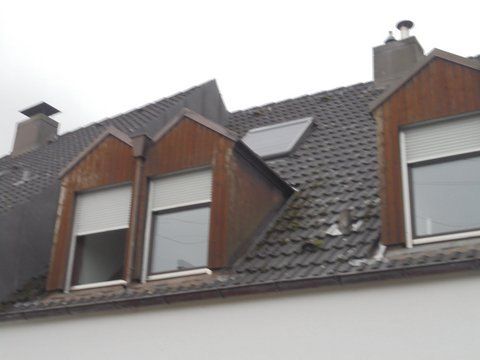 Ein Wohnhausdach mit Ziegeldeckung, drei Dachgauben mit Holzverkleidung und zwei Schornsteinen vor grauem Himmel.