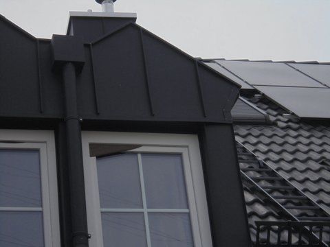Nahaufnahme eines mit schwarzem Metall verkleideten Dachgaubenfensters auf einem Ziegeldach; im Hintergrund sind Solarpaneele zu sehen.