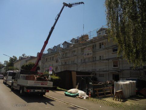 Eine Ansicht aus der Froschperspektive auf ein dreieckiges Giebelfenster mit dunklen Dachziegeln vor einem bewölkten blauen Himmel.