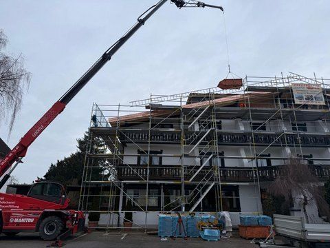 Ein roter Mobilkran hebt Baumaterialien auf das Dach eines mehrstöckigen, mit einem Gerüst bedeckten Gebäudes.