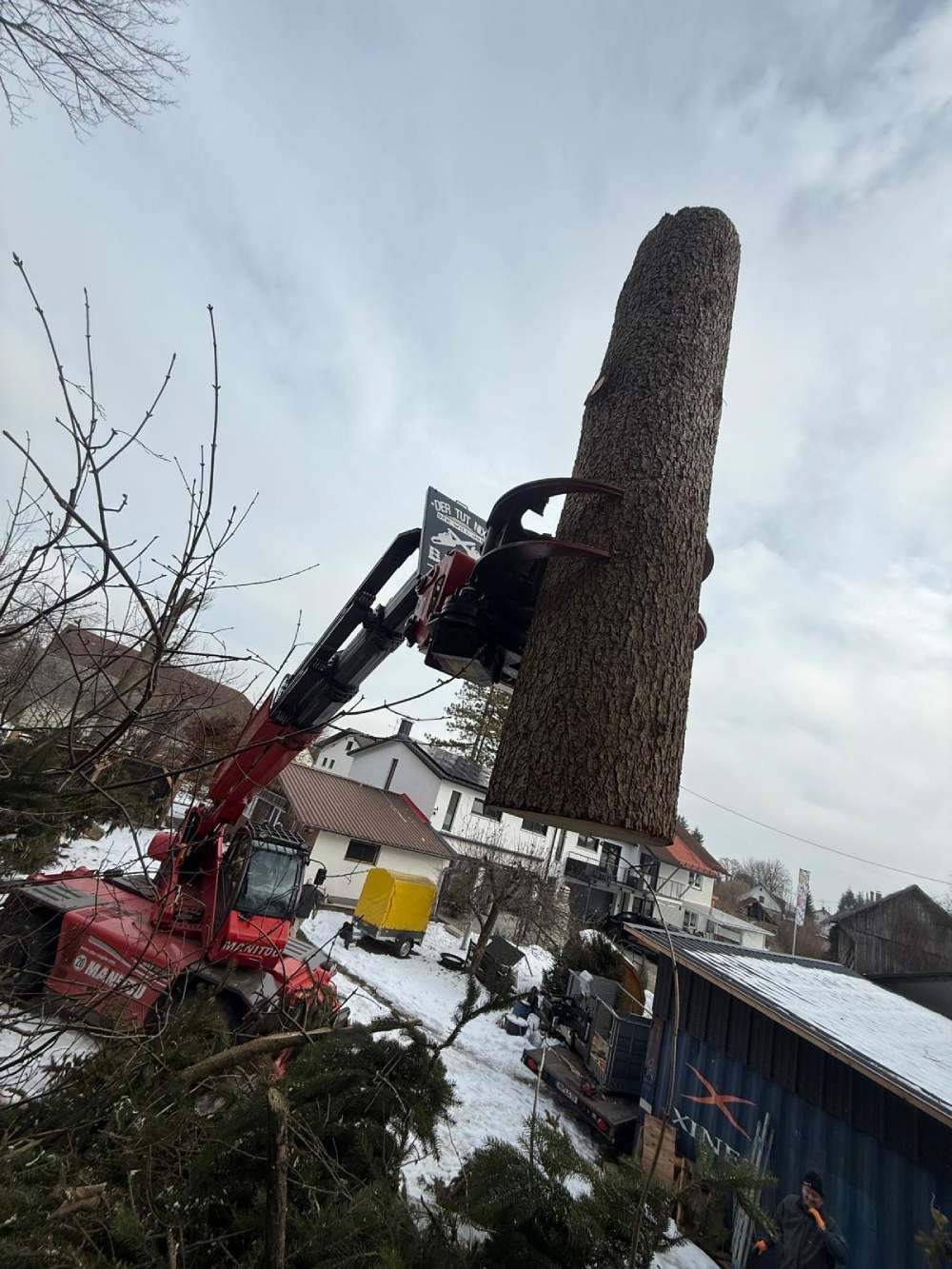Ein roter Kran hebt ein großes Baumstammsegment über ein verschneites Wohngebiet.