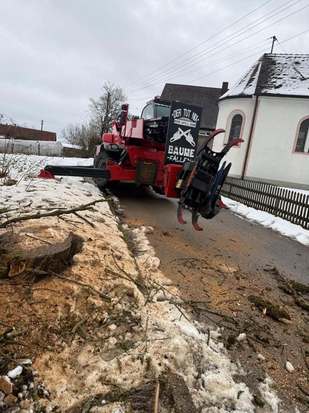 Eine rote Forstmaschine mit schwarzem Greiferprozessor parkt auf einer verschneiten Landstraße in der Nähe eines weißen Kirchengebäudes.