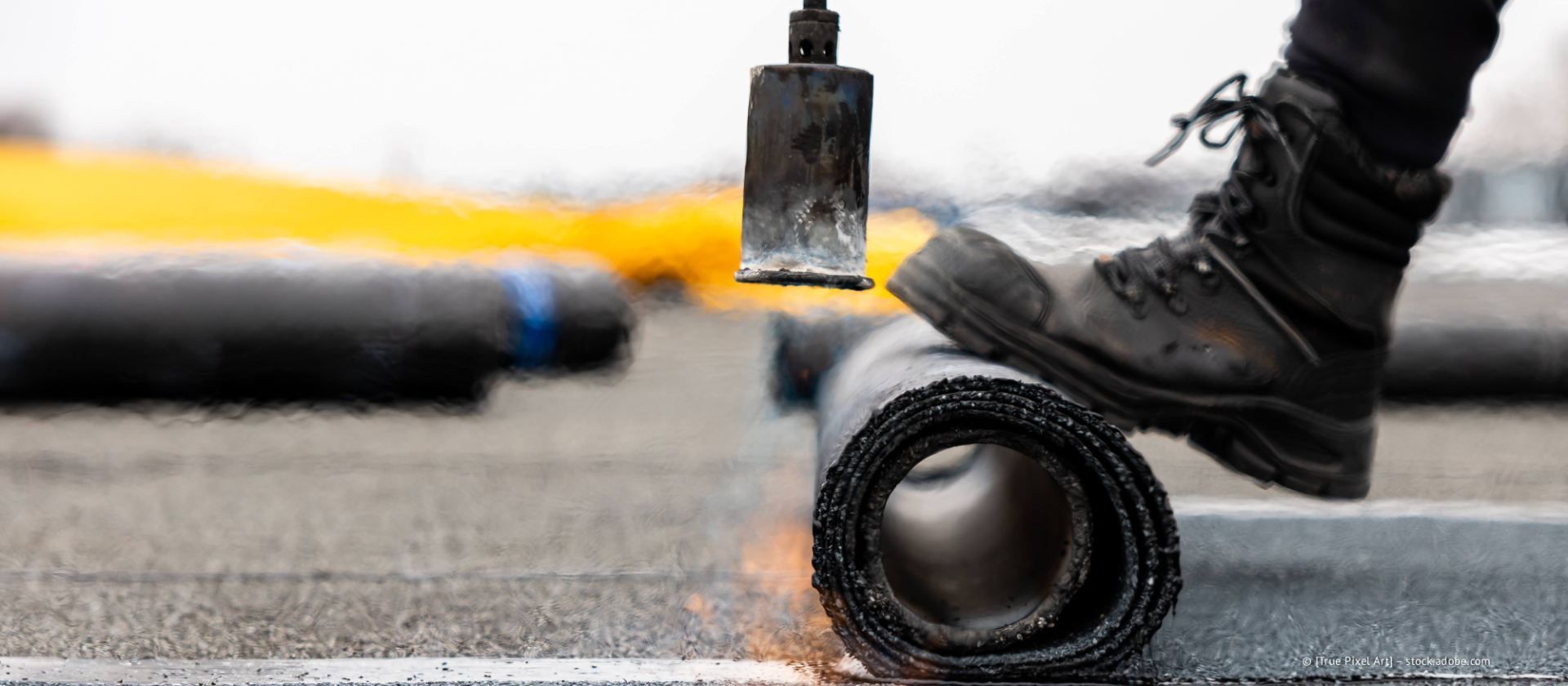 Ein Arbeiter drückt mit dem Stiefel eine Rolle schwarzes Dachmaterial zusammen, während ein Propangasbrenner die Oberfläche für die Montage anschmilzt.