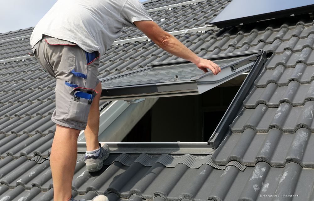 Eine Person in grauer Arbeitskleidung steht auf einem Ziegeldach und öffnet ein Dachfenster.
