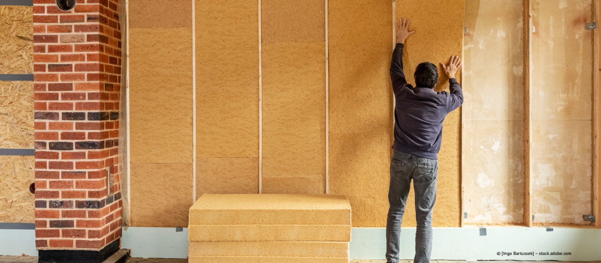 Eine Person montiert rechteckige Dämmplatten auf den Holzständern einer unfertigen Wand neben einer gemauerten Säule.