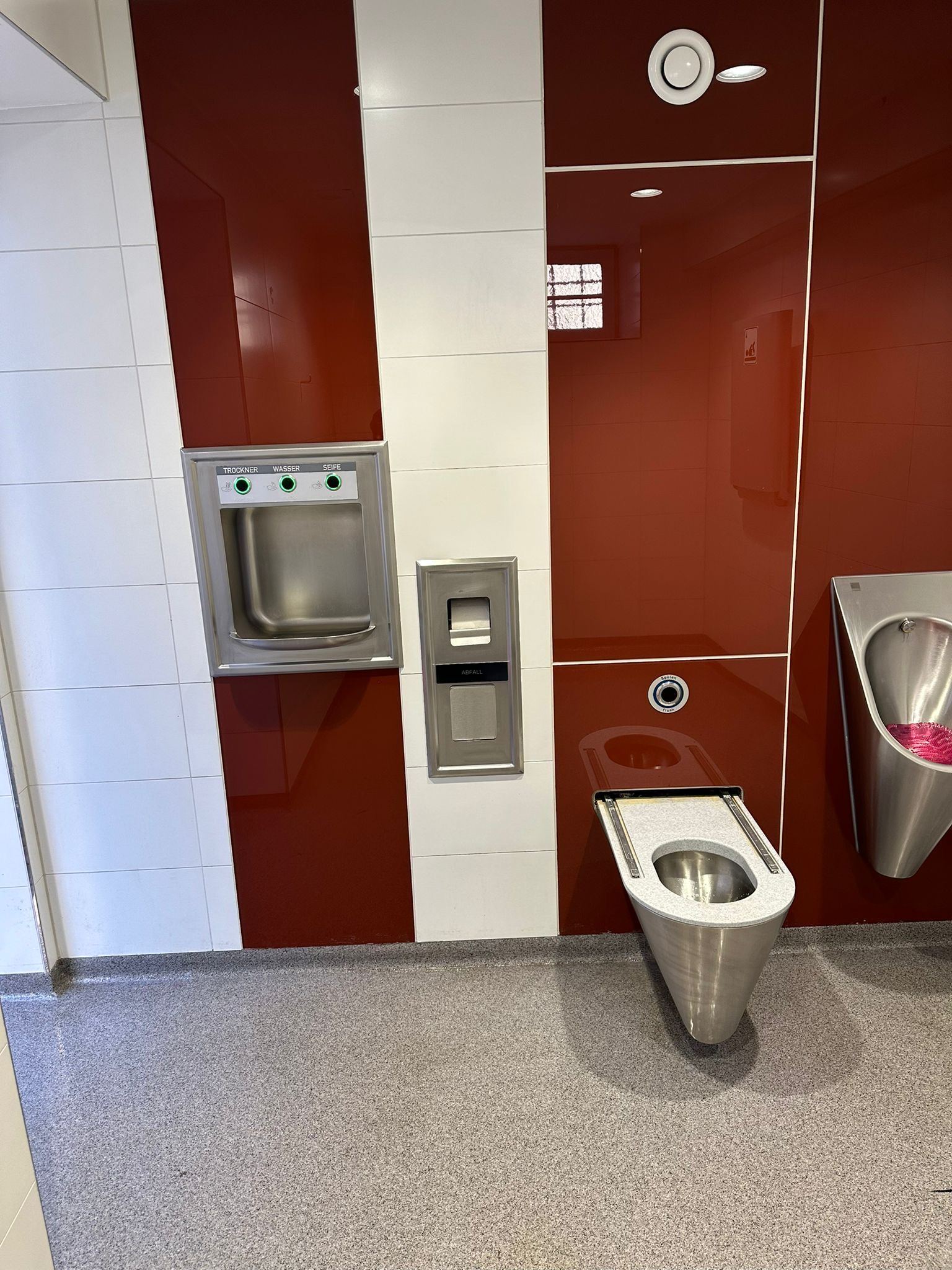 Waschbereich einer öffentlichen Toilette mit Händetrockner und Edelstahl-Waschbecken.