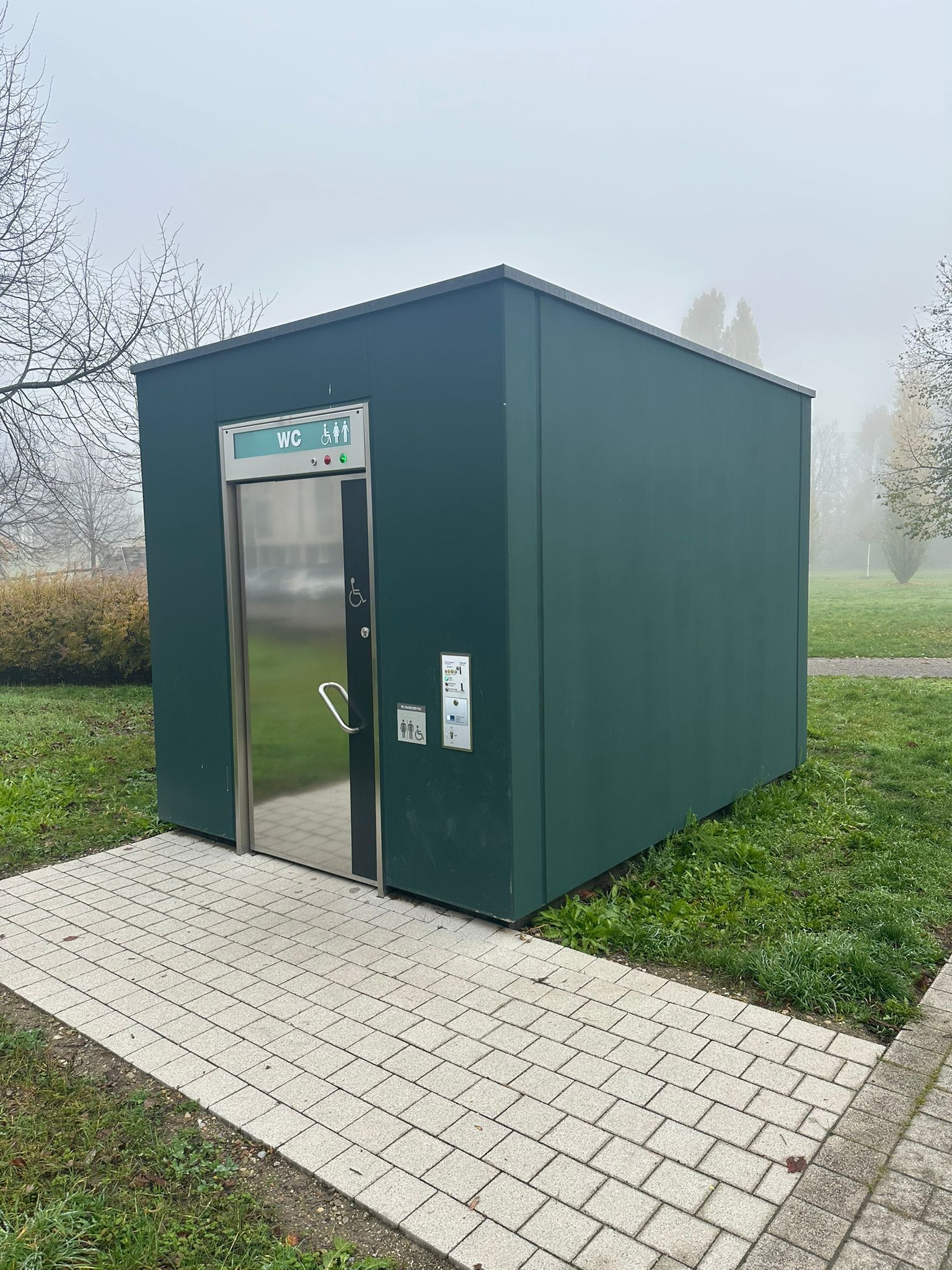 Außenansicht einer frei stehenden, grünen öffentlichen Toilettenanlage mit Edelstahltür.