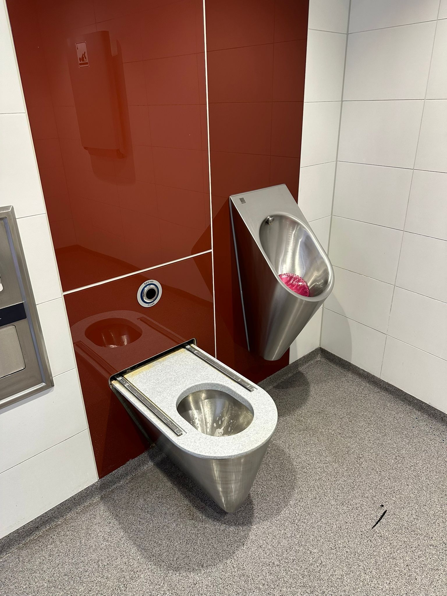 Urinal und Edelstahl-WC in einer öffentlichen Toilette mit rot-weißen Wandpaneelen.