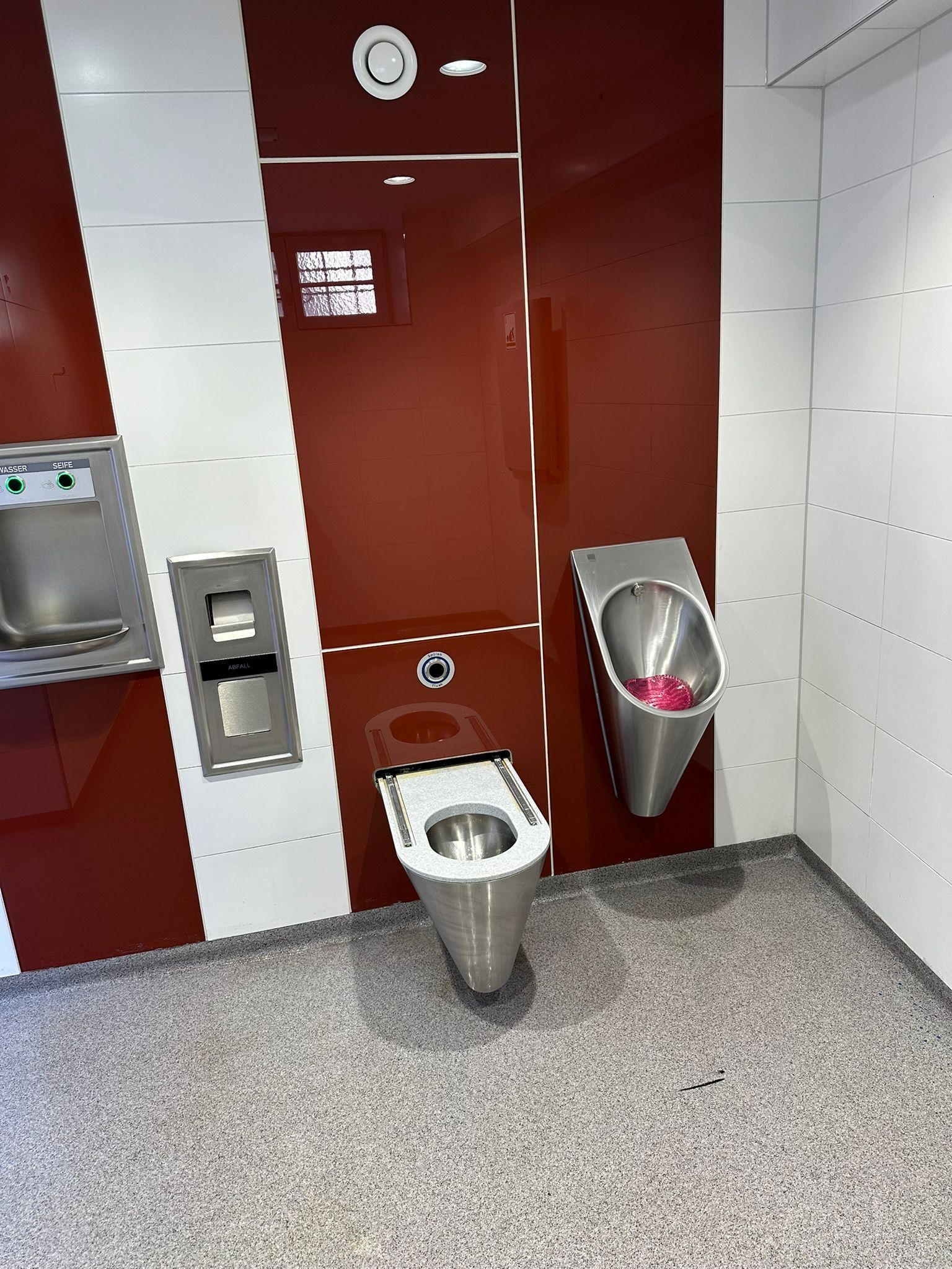 Innenansicht einer Sanitäranlage mit Urinal und Edelstahl-WC vor rot-weißen Wandpaneelen.