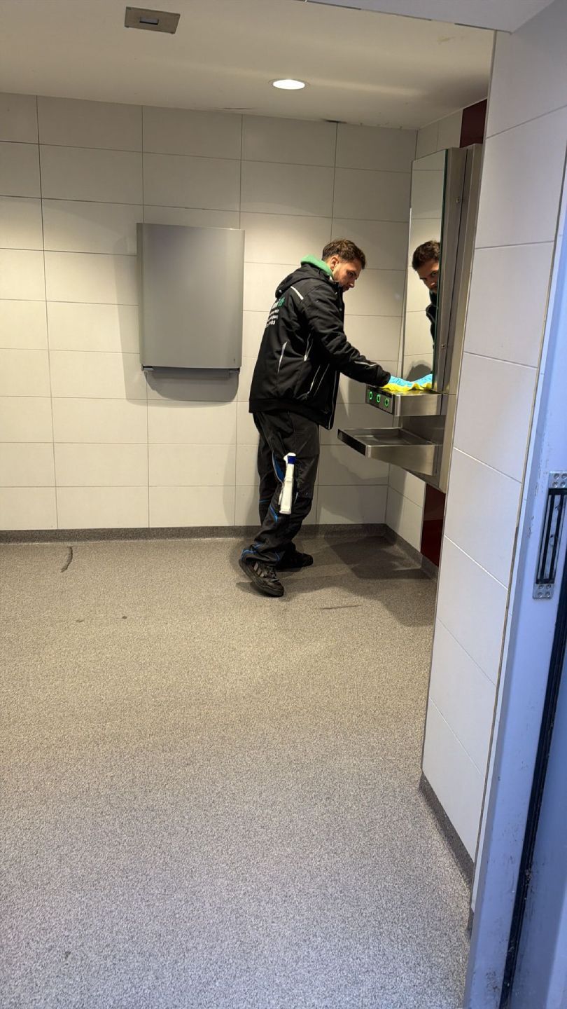 Innenraum einer öffentlichen Toilette mit Waschbecken, Seifenspender und einer Person, die sich die Hände wäscht.