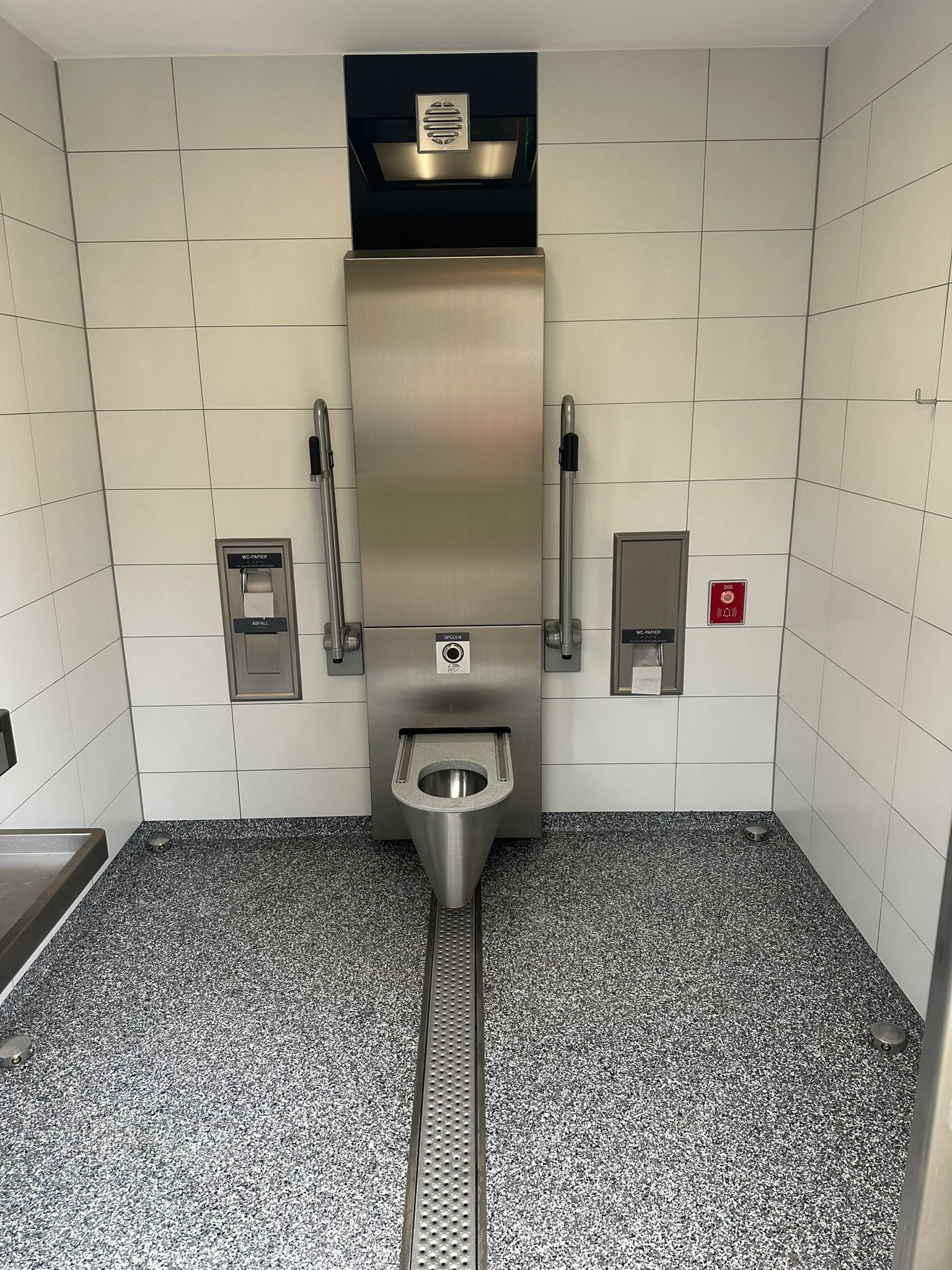 Innenraum einer behindertengerechten Toilette mit Edelstahl-WC und Haltegriffen.