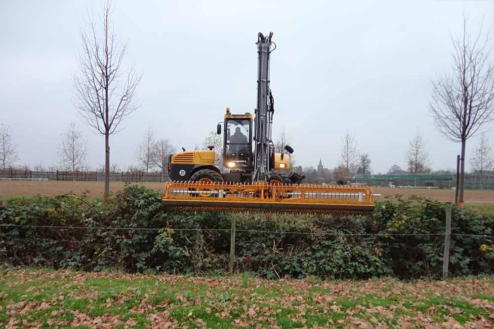 Ein gelber Traktor schneidet eine Hecke auf einem Feld.